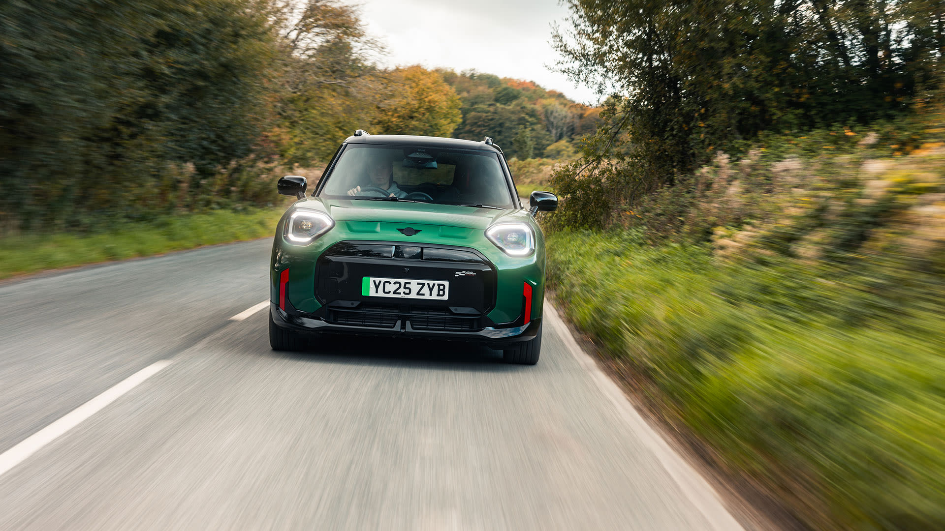 Green MINI Aceman driving along a tree-lined country road.