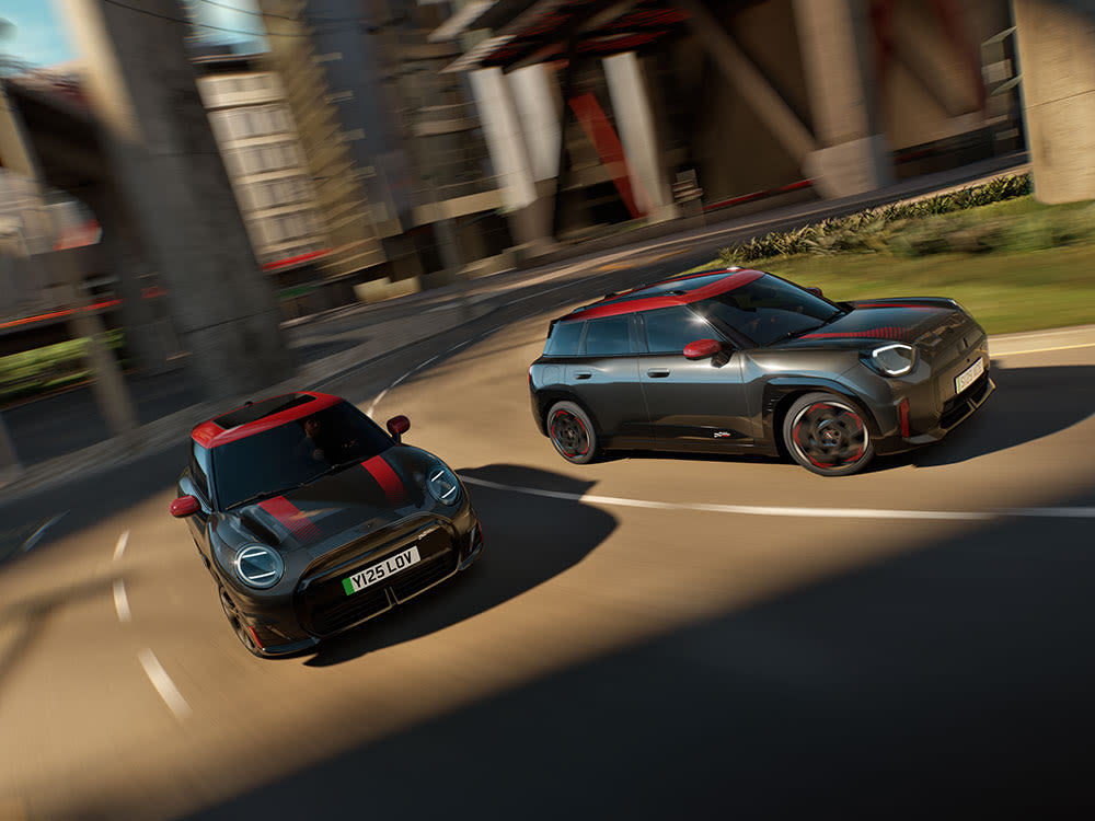Two MINI cars with red roof accents driving on a city road, captured with motion blur against a modern architectural background.