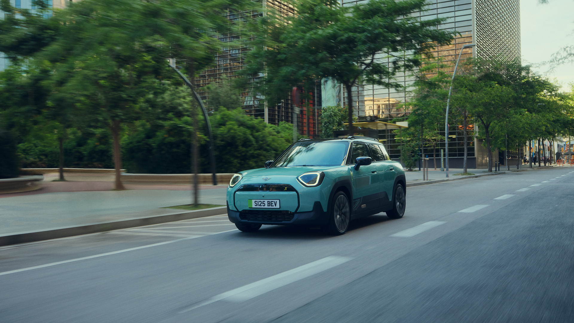 Front-left view of a tourquase MINI Aceman driving on a public city road with high buildings and trees in the background