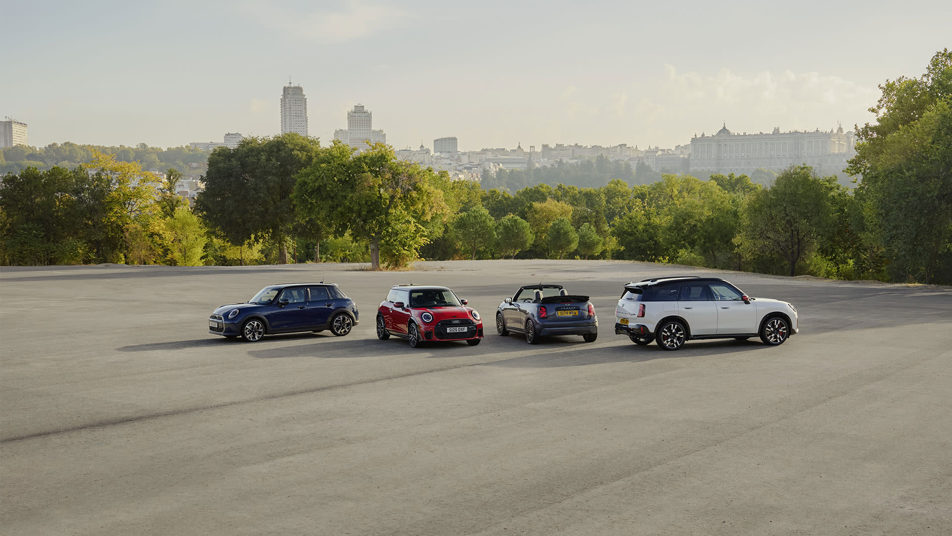 Four MINI cars parked in an open area with trees and a city skyline in the background.