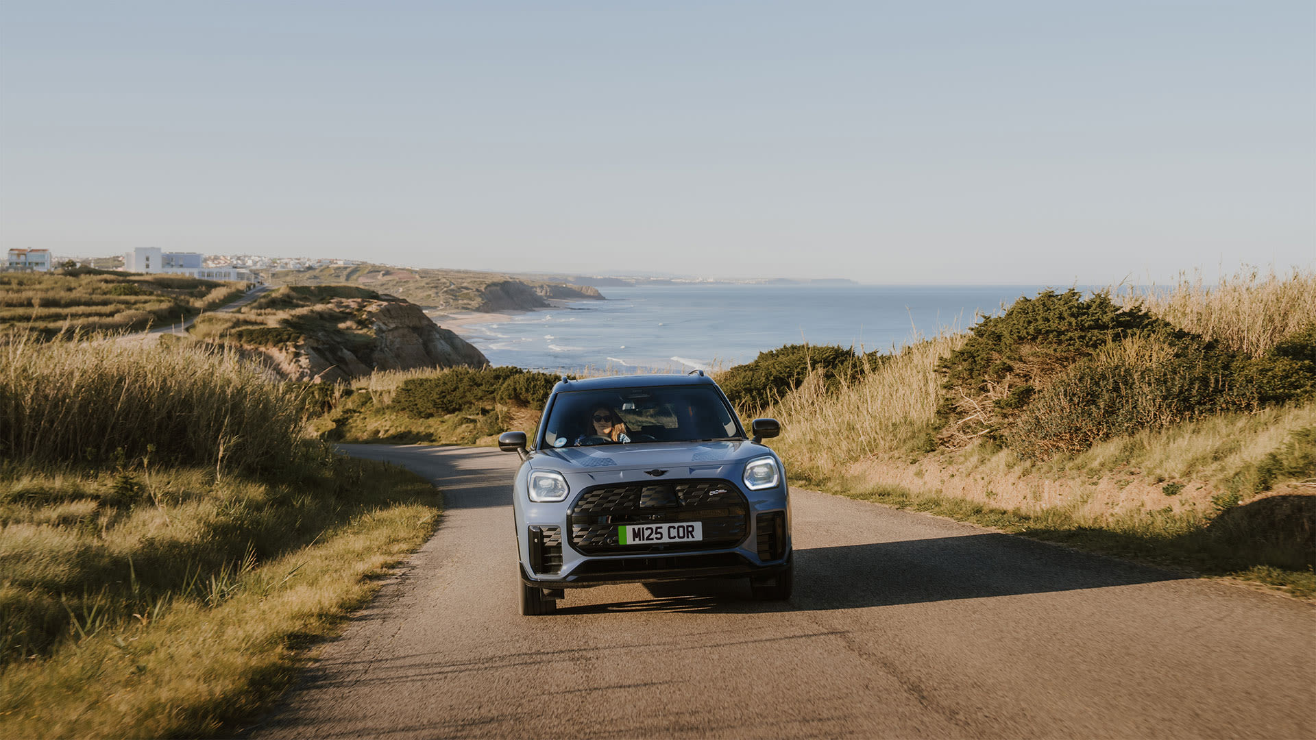 Blue MINI Countryman driving along a coastal road with the sea and cliffs in the background.