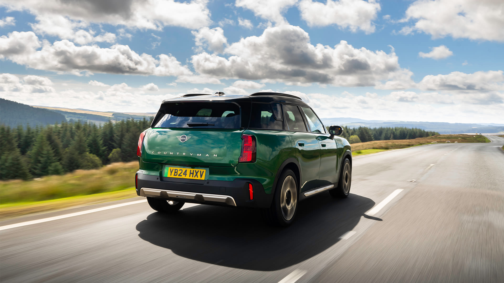 Green MINI Countryman driving on a scenic road with trees and hills in the background under a partly cloudy sky.