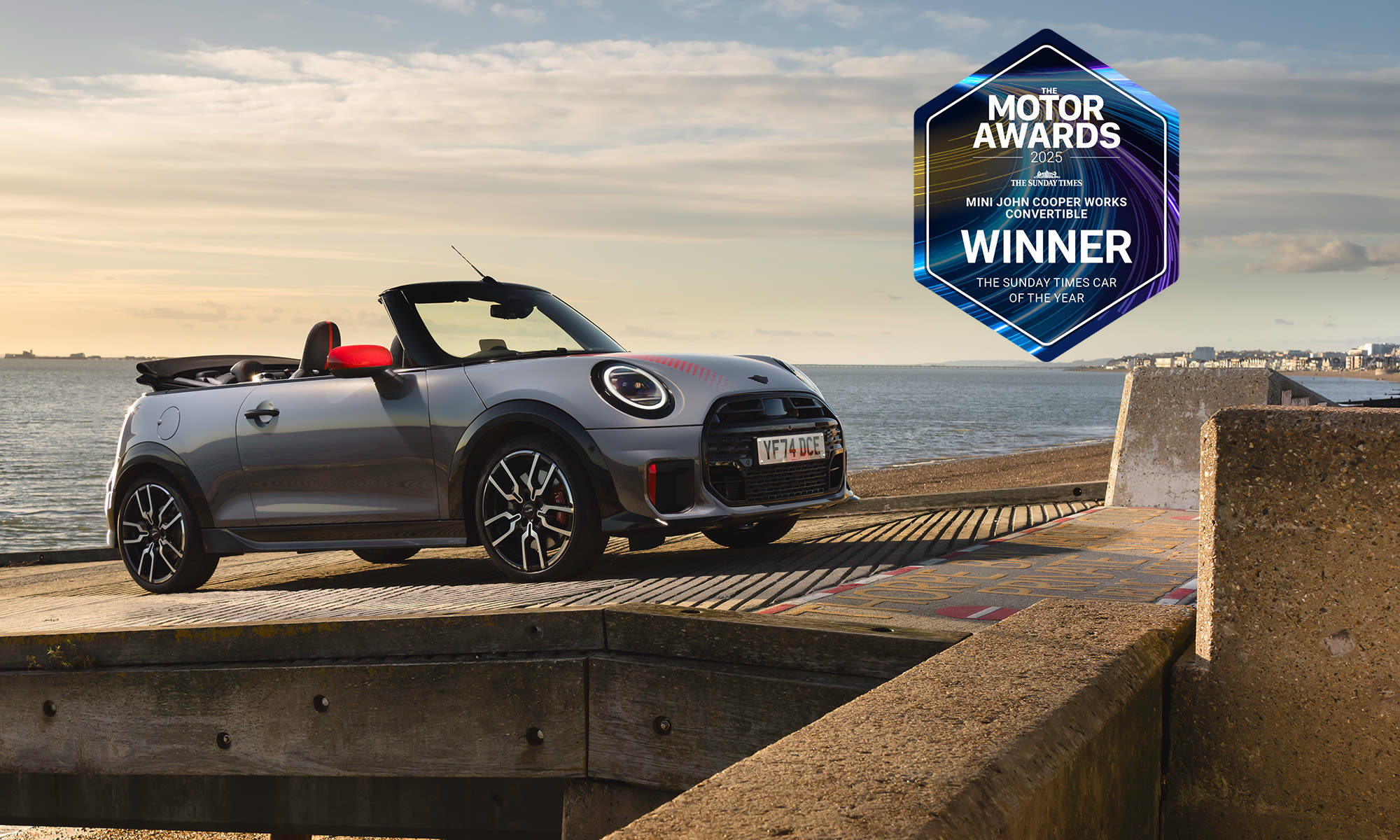 Side view of a MINI JCW Convertible parked on a seaside promenade, shown with a motor awards winner badge.