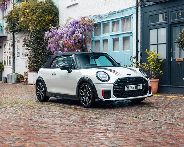 White MINI Convertible with black soft top parked on a cobbled street lined with colourful houses and flowering plants.