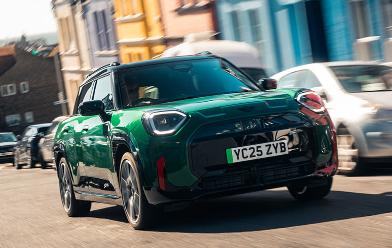 Front view of a green MINI Aceman JCW driving on a city road with cars parked on the left side