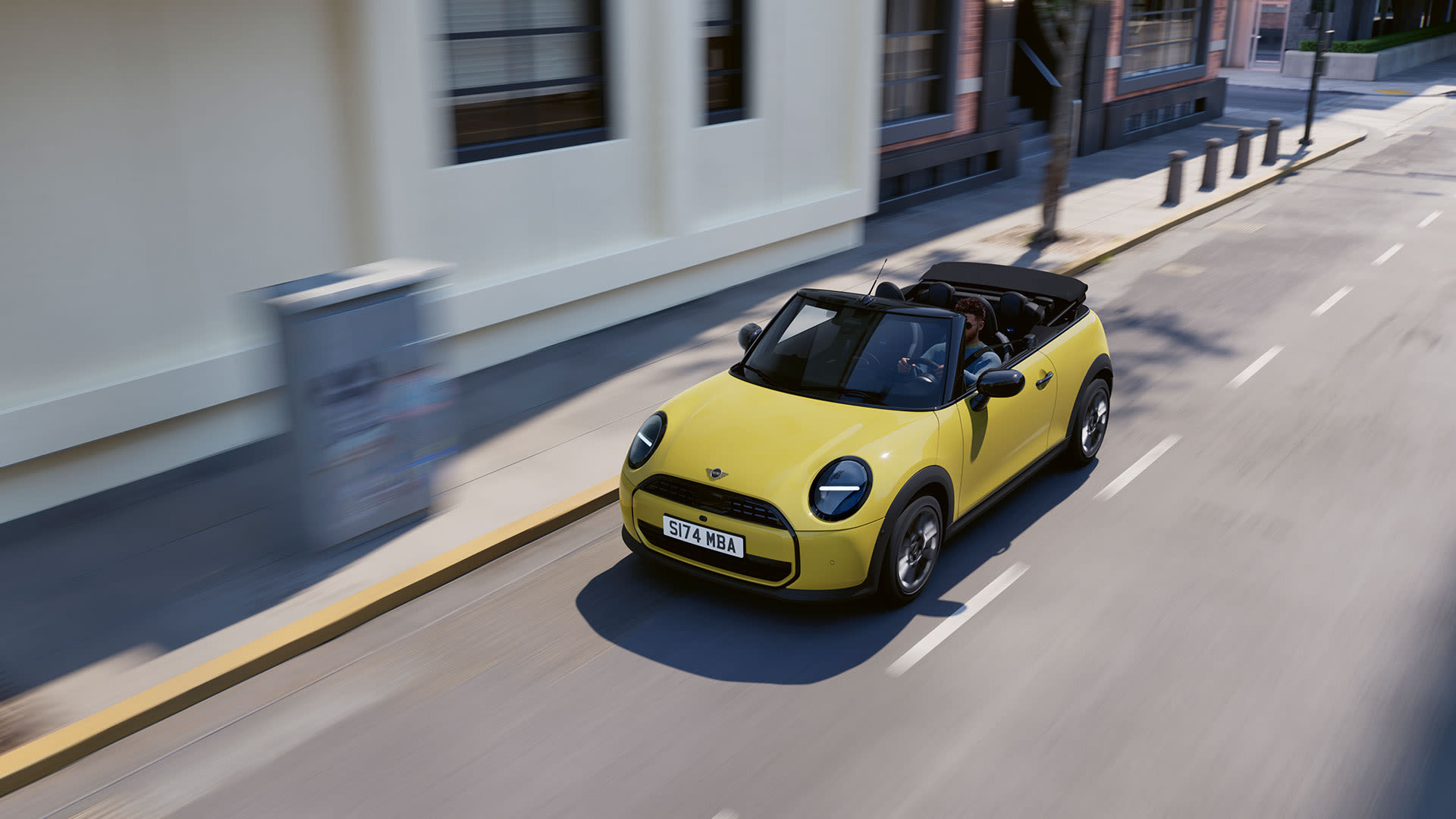 Yellow MINI Convertible driving along a city street with the roof down, shown from an elevated angle.