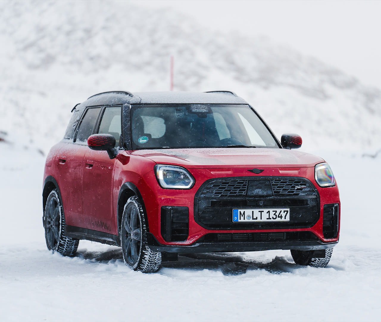 Red MINI SUV with black roof and wheels, parked on a snowy landscape with snow-covered mountains in the background.