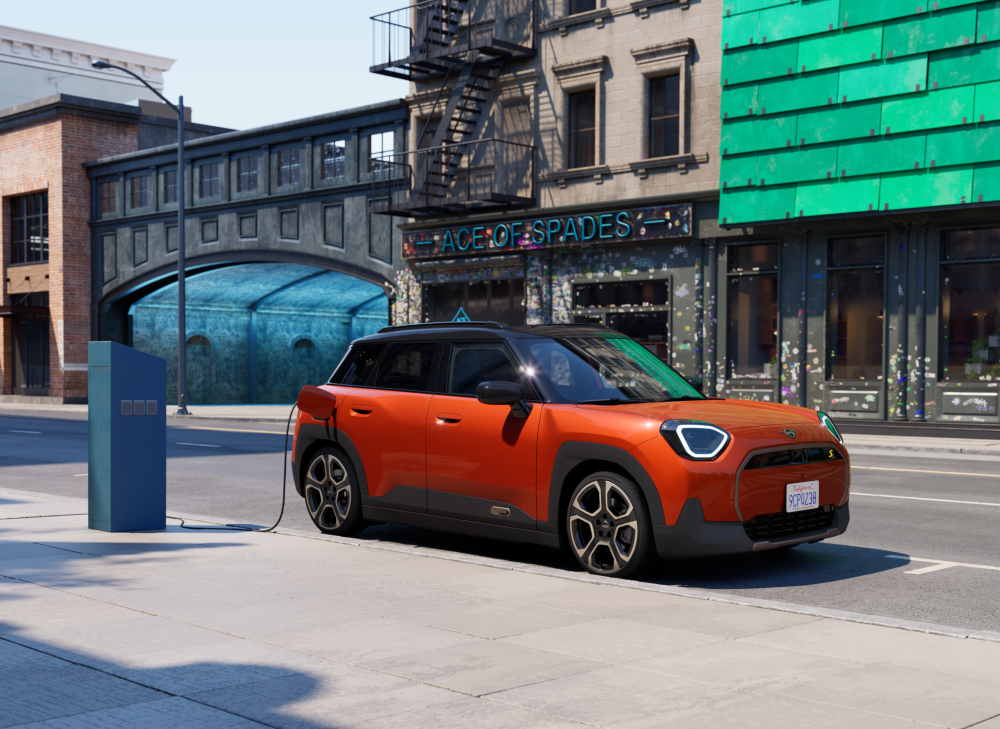 Orange MINI Aceman charging at a street-side charging station in an urban area with buildings in the background.