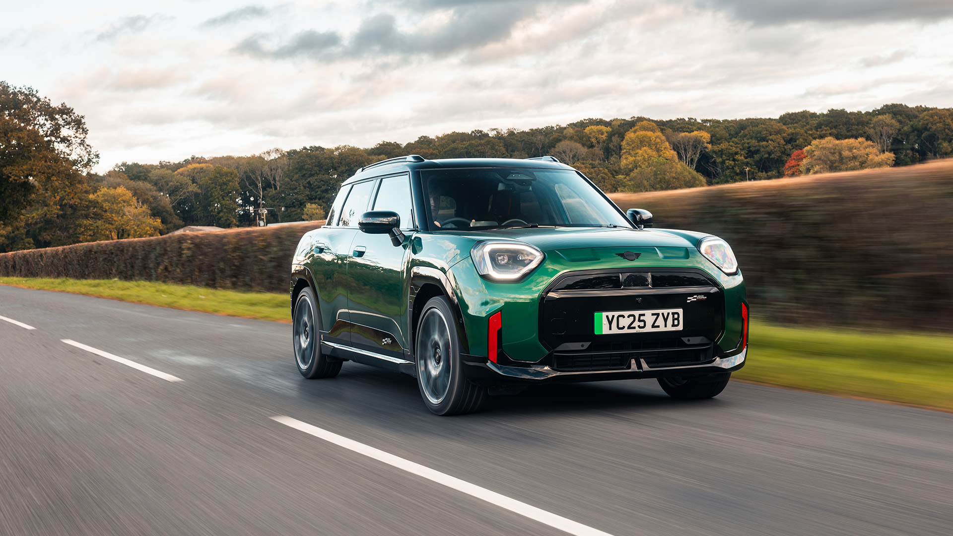 Front right side view of a green MINI Aceman JCW Electric, driving on a country road with hedges on the side