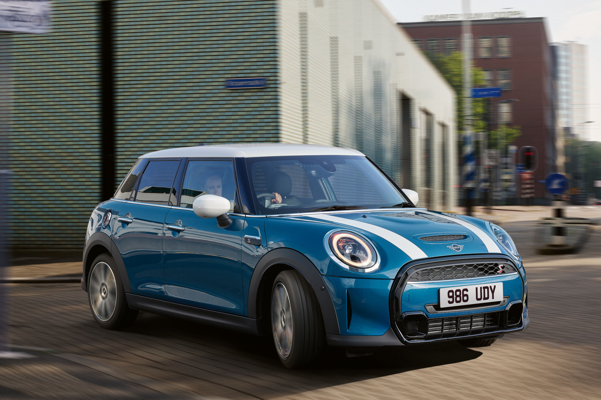 Blue MINI 5‑Door Hatch driving through a city street, shown in motion with an urban background.