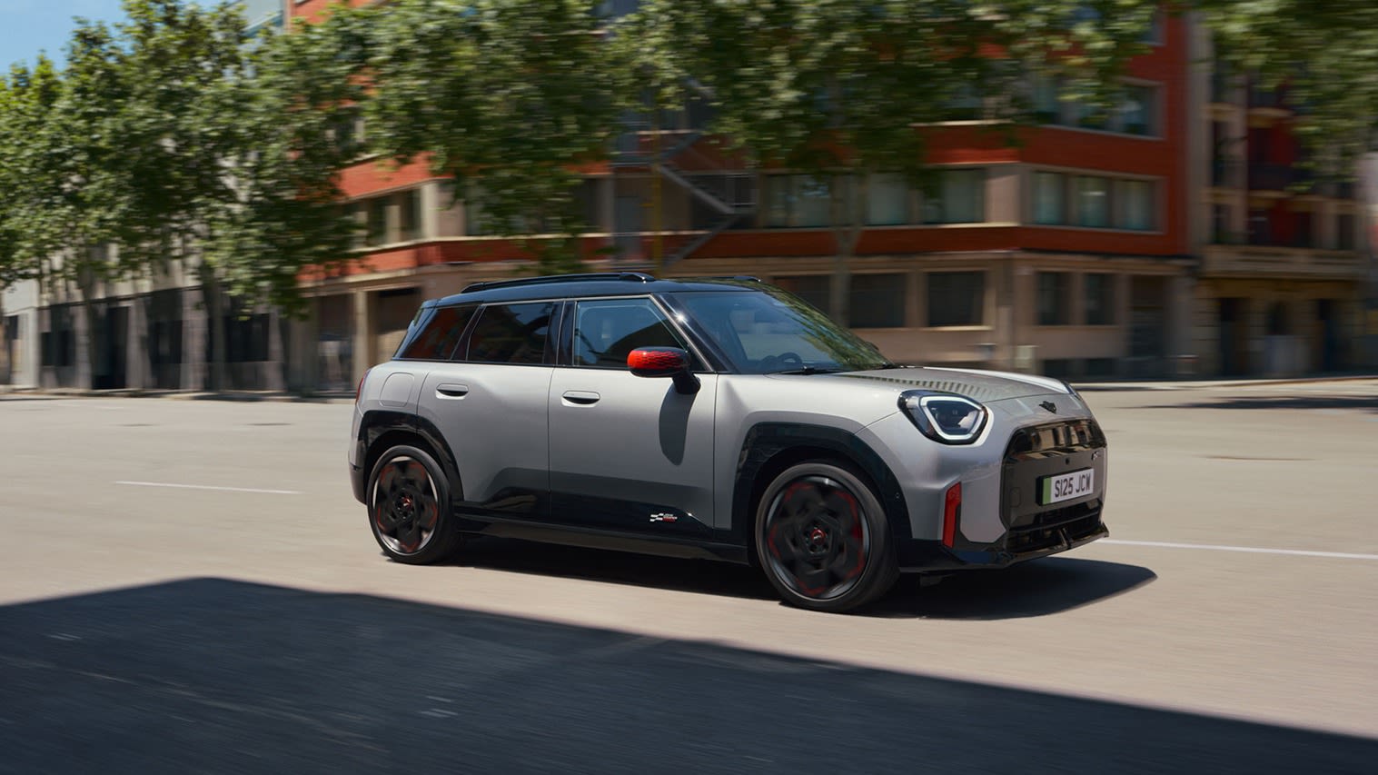 Silver MINI Aceman driving on an empty urban street with a modern building and trees in the background.