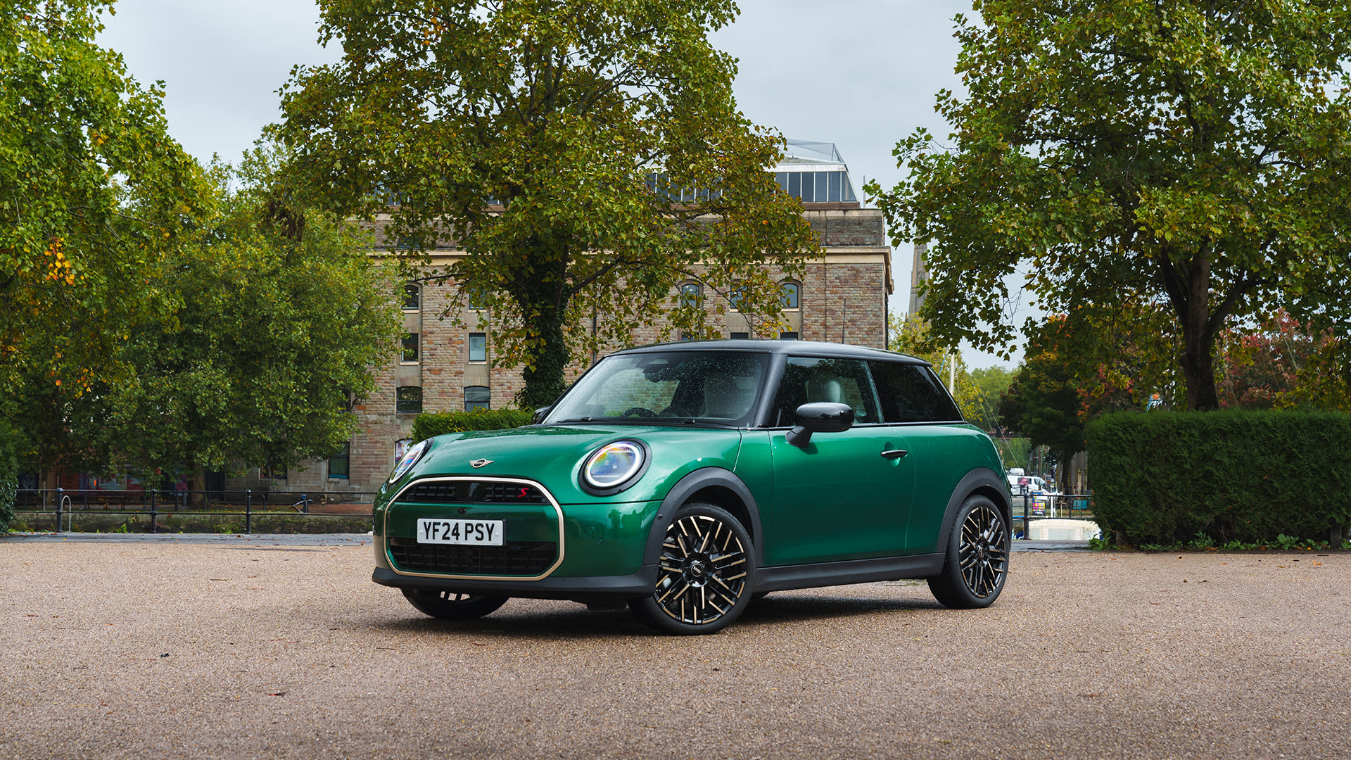 Green MINI Cooper S hatchback parked on a tree-lined drive, with a historic stone building in the background.