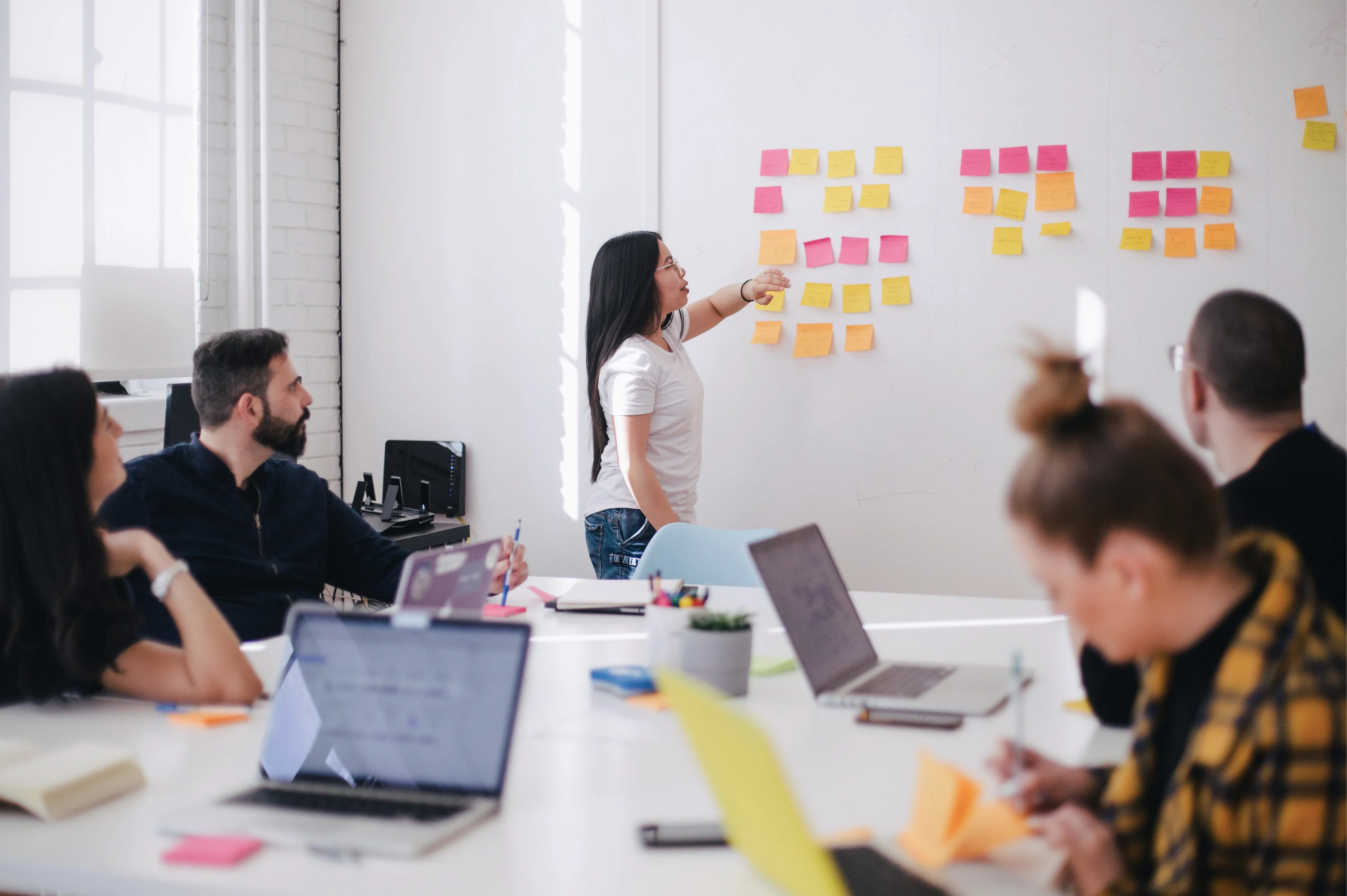 A table of people in front of a woman standing in front of a white wall of sticky notes