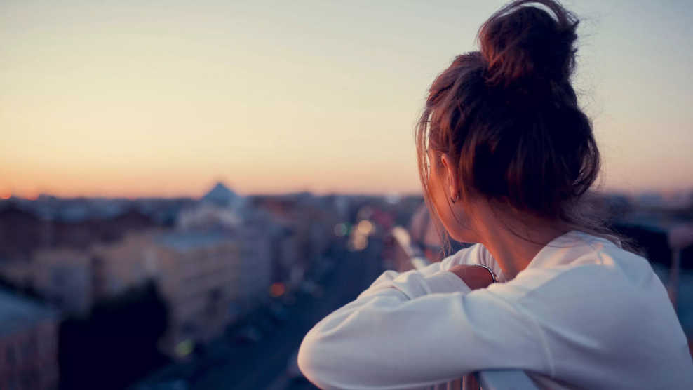 eine Frau mit zugemachten Haar und langen weißen Shirt bei Abendrot von einem Balkon über die Dächer einer die Stadt blickt