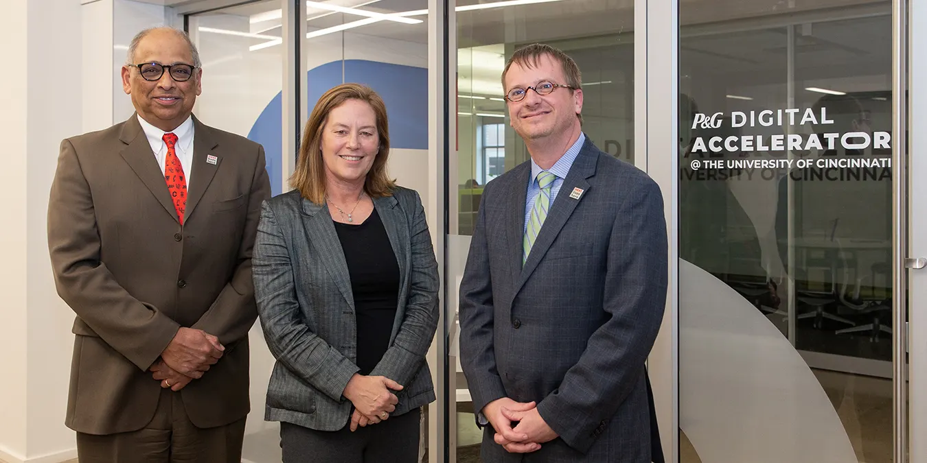 Three persons standing looking towards the camera, wearing business suits. Text at the bottom: P&G Digital Accelerator @ The University of Cincinnati Logo