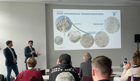 Two presenters stand at the front of a conference room giving a presentation on post-household transformation, with a projected slide showing stages such as sorting, washing, and Flexloop processing.