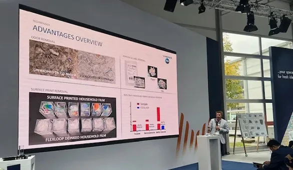 A presenter speaking onstage in front of a large screen displaying an overview of technology advantages, including odor removal, surface print removal, and sample data comparisons.