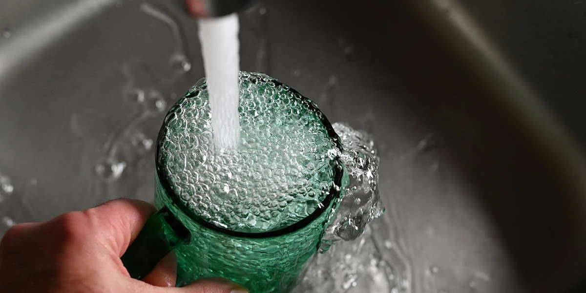 A hand holding a green glass cup being filled with water from a tap, with water overflowing