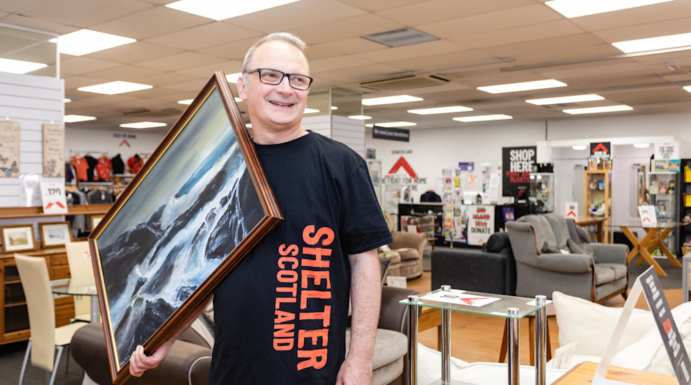 A white male furniture shop volunteer smiles at the camera. He is wearing a Shelter Scotland t-shirt and carrying a painting, which is a dramatic ocean scene in a wooden frame. 