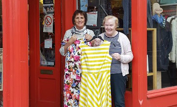 Two ladies hold up patterned dresses outside a Shelter Scotland shop.