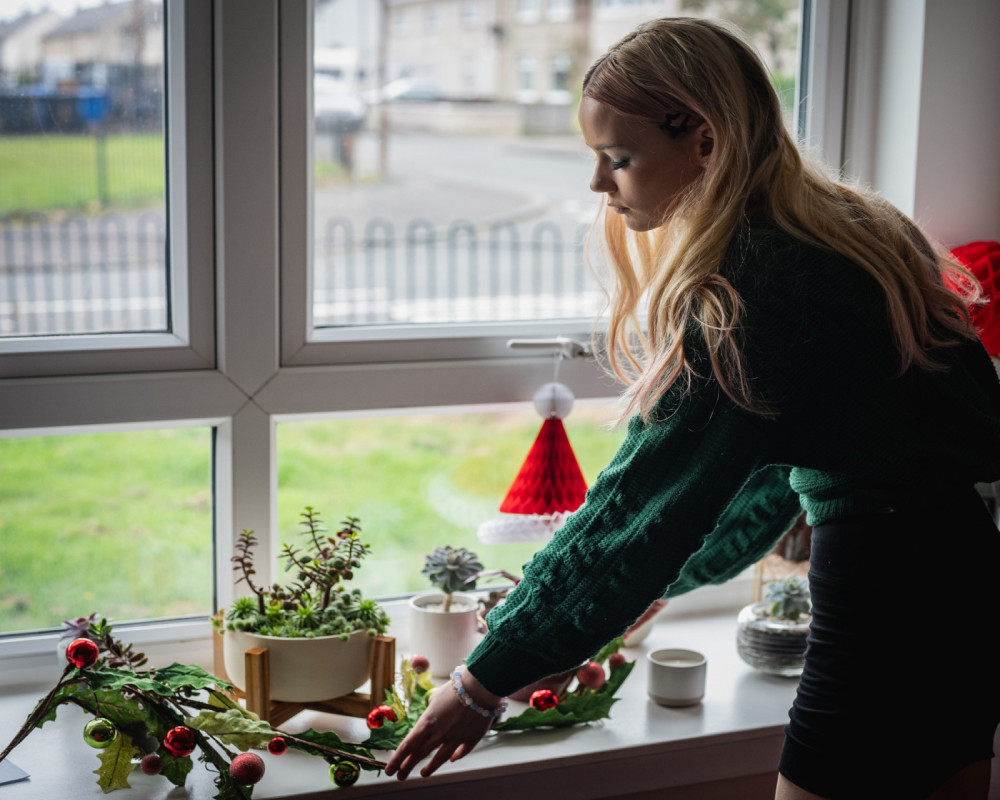 B stands at her window, there is a view of the street outside. The window sill has Christmas decorations.