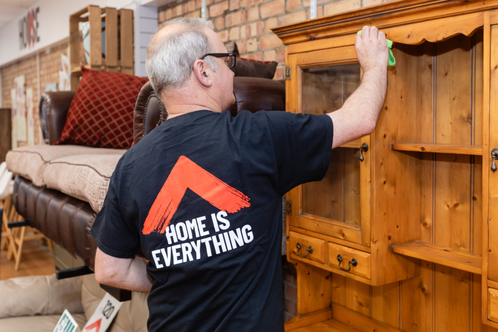 A white male furniture shop volunteer dusts a wooden cabinet for sale. He is wearing a Shelter t-shirt, the back reads 'Home is everything'.