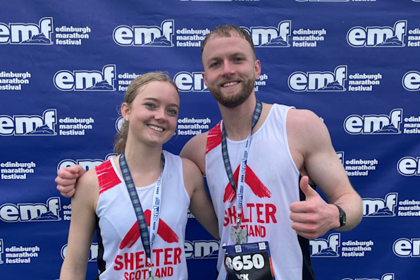 Two smiling runners celebrate after their race, wearing medals and Shelter Scotland running vests. The backdrop has the Edinburgh Marathon Festival logo.