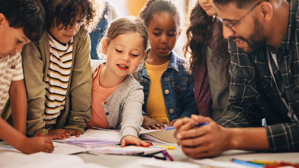 Young children around a table with their teacher, choosing from lots of reading material