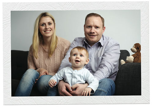 A mother, father and baby sit on a couch together, all smiling