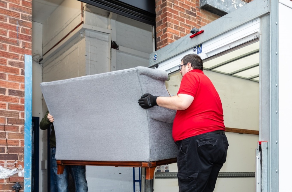 Two Shelter Scotland furniture shop volunteers carry a grey sofa off the collection van and into the delivery entrance.