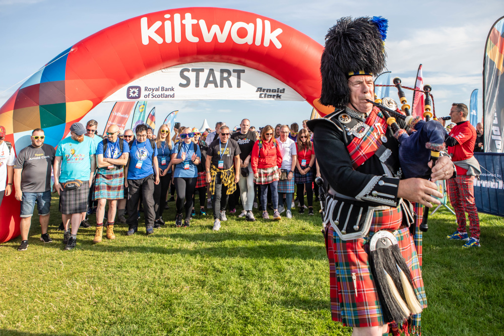 People stand under the Kiltwalk banner, ready to start their walk. There is a bagpiper in the foreground.