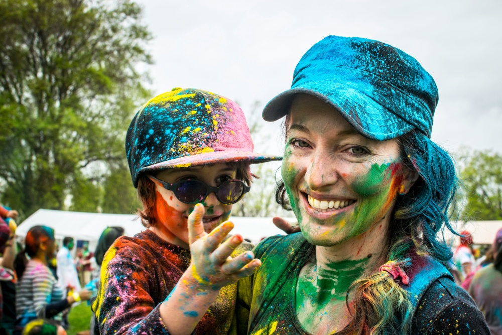 A woman wearing a blue hat holds a child wearing sunglasses and a multicoloured hat. Both are covered in colourful powder.