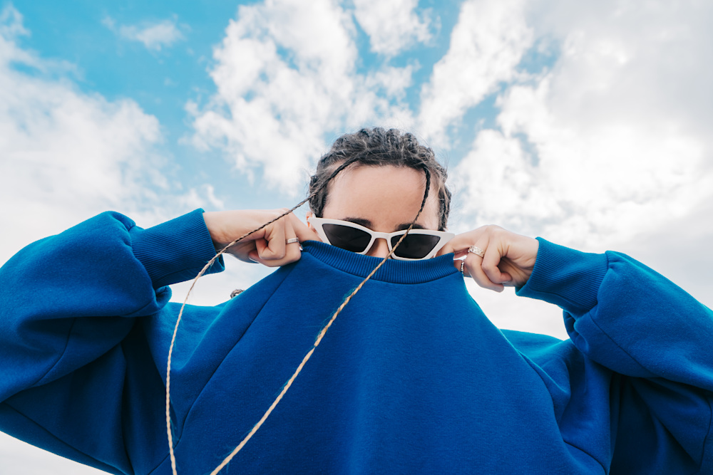 A woman in a blue jumper and sunglasses stands outside while pulling her top up over her mouth