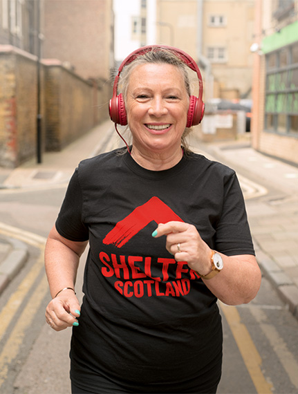 A woman wearing headphones and a T-shirt with a Shelter Scotland logo is smiling as she runs down a city street