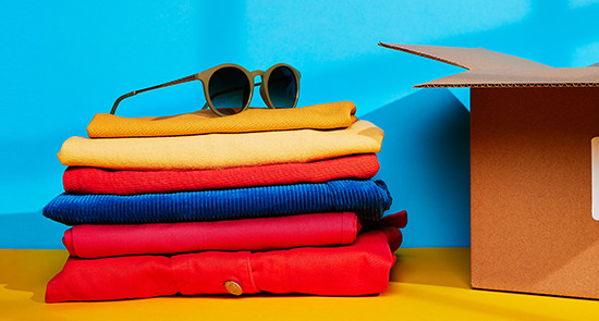 A folded pile of colourful donated clothes, with a pair of sunglasses siting on top, next to a cardboard box, from a collection of items donated to Shelter