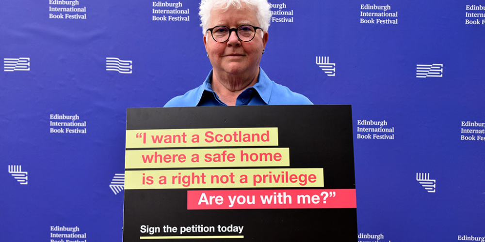 Val McDermid pictured at Edinburgh International Book Festival holding a sign that says: "I want a Scotland where a safe home is a right not a privilege. Are you with me?" Sign the petition today.