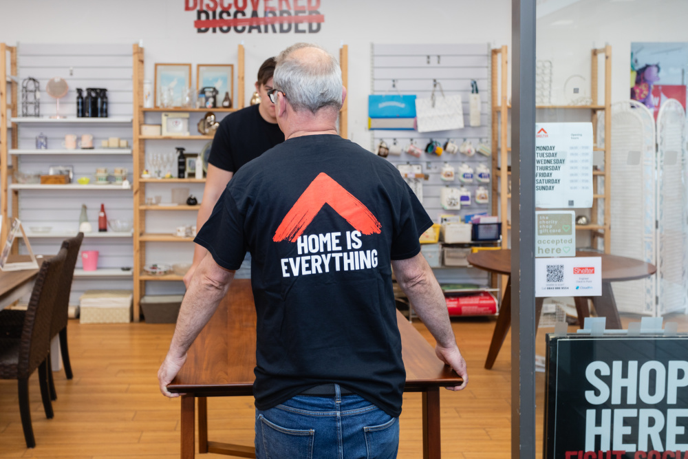 Two white male volunteers, one older and one younger, carry a wooden coffee table into the furniture shop. One faces away from the camera, and the back of his t-shirt reads 'Home is everything'.