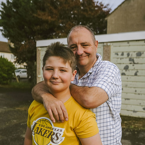 Man in checked shirt with arms around son's shoulders, boy is wearing yellow t shirt with Lakers logo. Both are smiling.