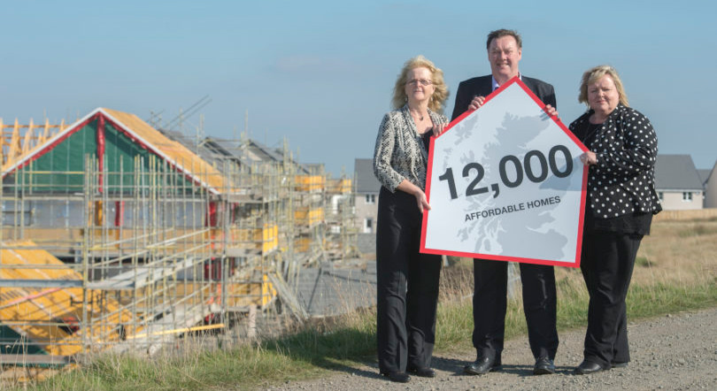 Houses being built in the background. Three people stand in front hold a sign with a Scotland map, which reads '12,000 affordable homes'.