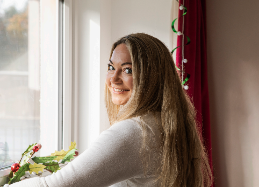 Grace stands at her window, which ahs Christmas decorations. She is smiling.