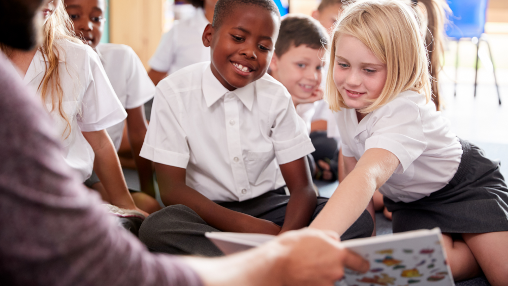 A group of pupils looking at a book being held by the teacher, one child is pointing at the book