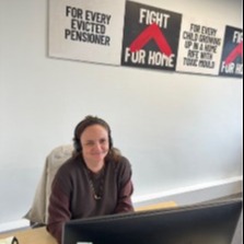 Jo, a woman with brown hair, is sitting behind a desk with a computer screen in front of her. She is wearing a brown cardigan and is smiling.
