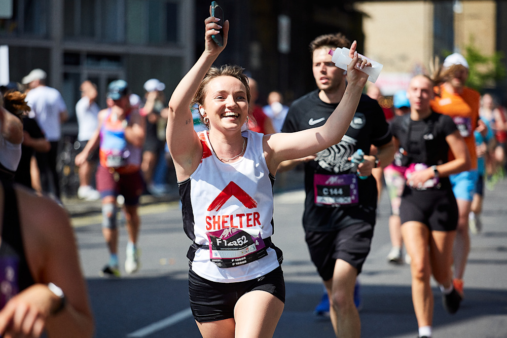 A woman in a Shelter vest runs past the cheer point of the half marathon, smiling and waving.