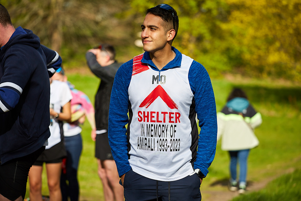 A male runner wearing a Shelter vest waits in a park before the race starts, smiling with his hands in his pockets.