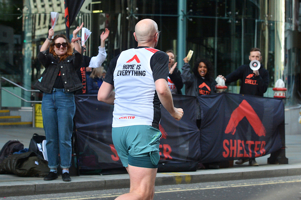 A man in a Shelter vest that reads 'Home is everything' on the back runs past the cheer point. People wearing Shelter hoodies excitedly wave, clap and shout through a megaphone.