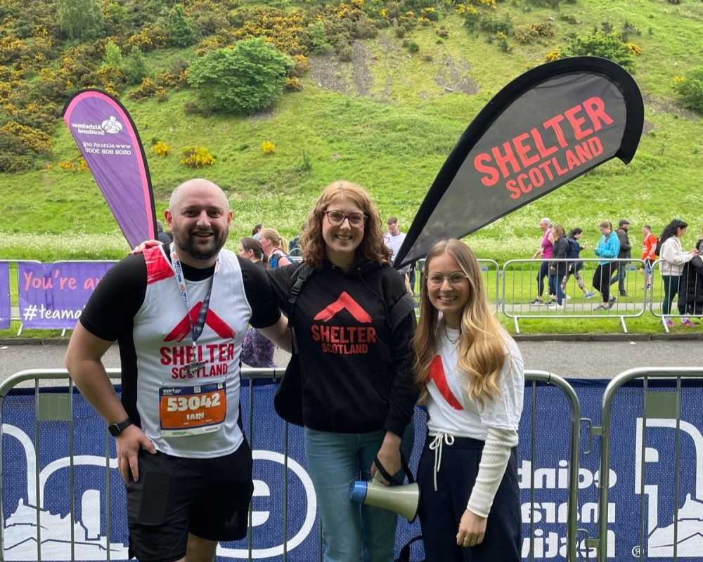 A marathon finisher wearing a Shelter Scotland vest and medal smiles at the camera next to two members of the Events team. In the background is a Shelter Scotland banner and green hilly scenery.