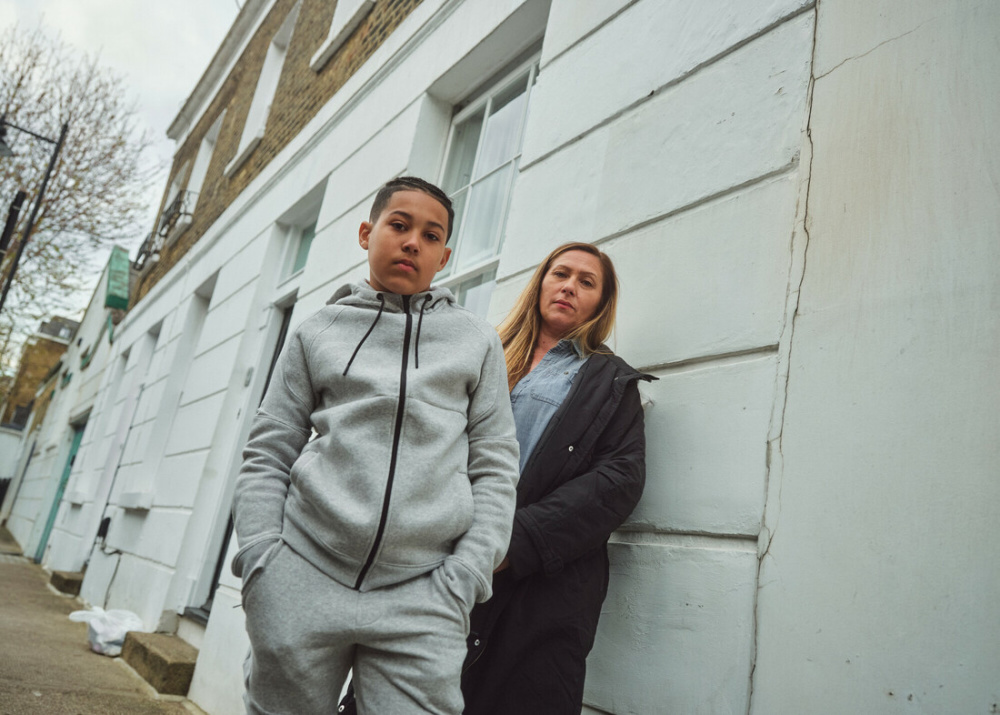 A mother leans on a wall whilst her son puts his hands in his pockets in a tracksuit outside in the street in London