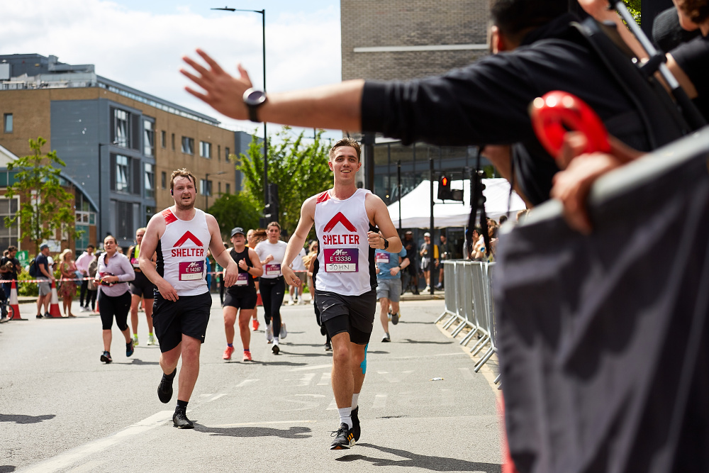 Two men in Shelter vests run towards the cheer point. A member of the Events team holds out his hand ready to give them a high five.
