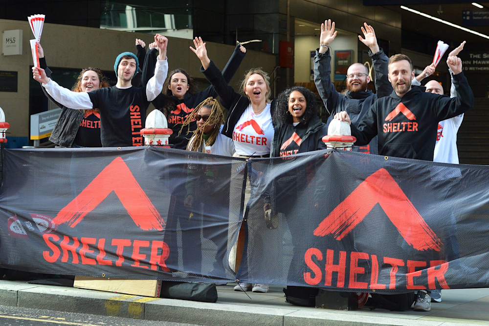 A group of Shelter staff and volunteers at the race cheer point, with big Shelter banners. They are wearing Shelter hoodies and waving at the camera.