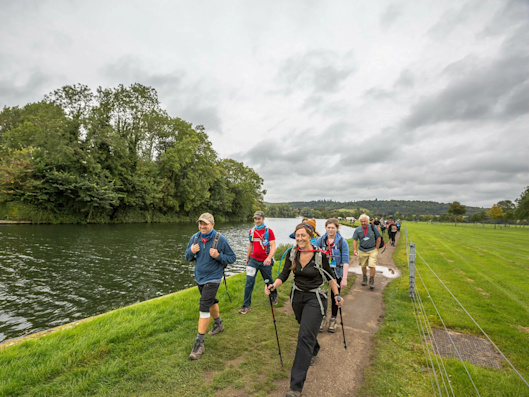 A group of Thames Path Challenge walkers with backpacks and hiking poles. They are hiking along the riverside with green fields and trees in the background.